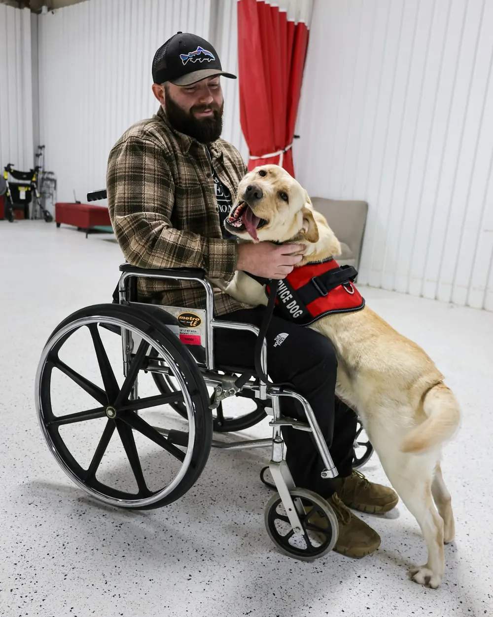 A TASDF recipient in a wheelchair greeting his yellow Labrador service dog in a red vest