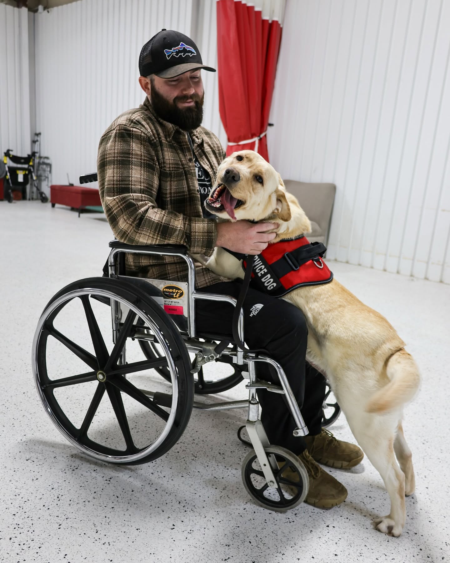 A TASDF recipient in a wheelchair greeting his yellow Labrador service dog in a red vest