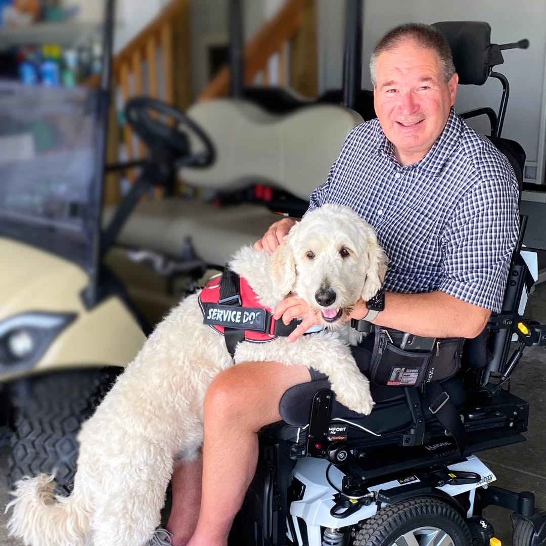 A man in a power wheelchair with his cream doodle service dog resting on his lap