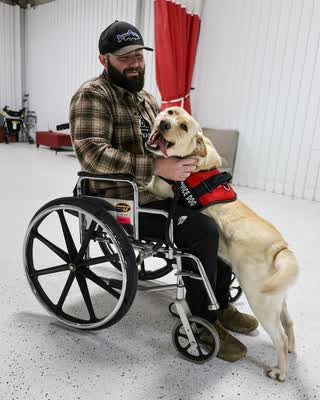 A bearded man in a wheelchair embracing his yellow Labrador service dog in a red SERVICE DOG vest