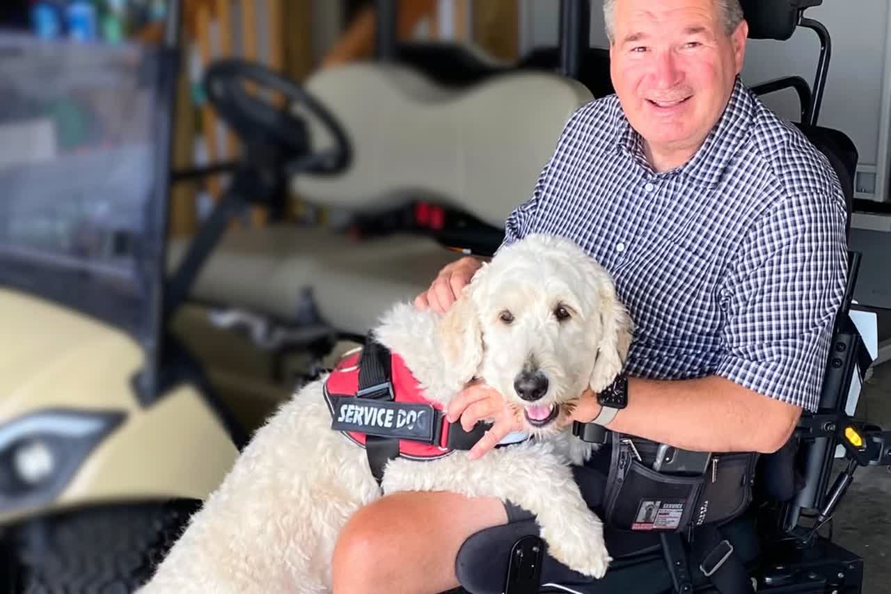 A smiling man in a power wheelchair with his white doodle service dog in a red SERVICE DOG vest